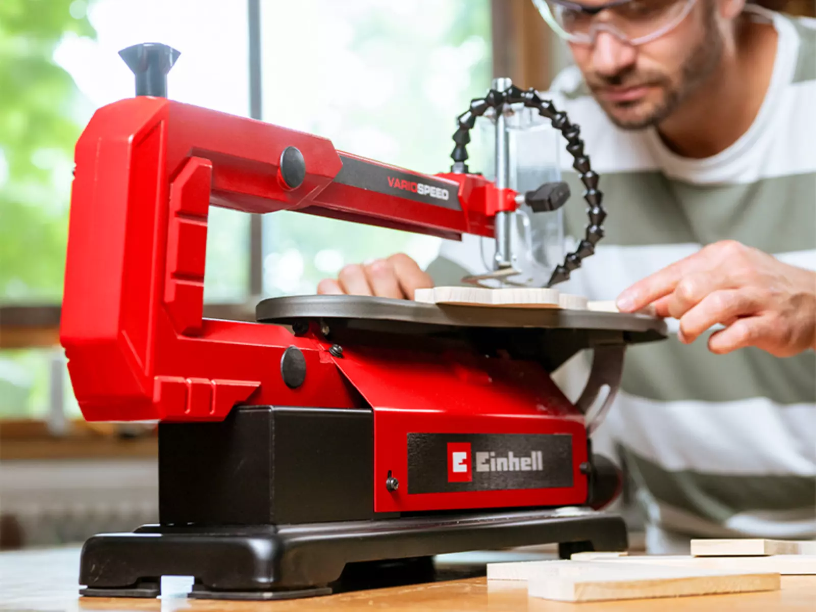 La sierra de marquetería TC-SS 406 E de Einhell se encuentra sobre una mesa; Un hombre con gafas protectoras corta un trozo de madera.