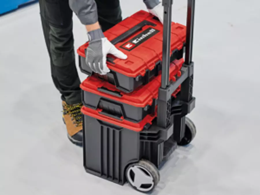 Angled view of a tool trolley with three stacked black-red Einhell E-cases. A person wearing gloves secures the top case.