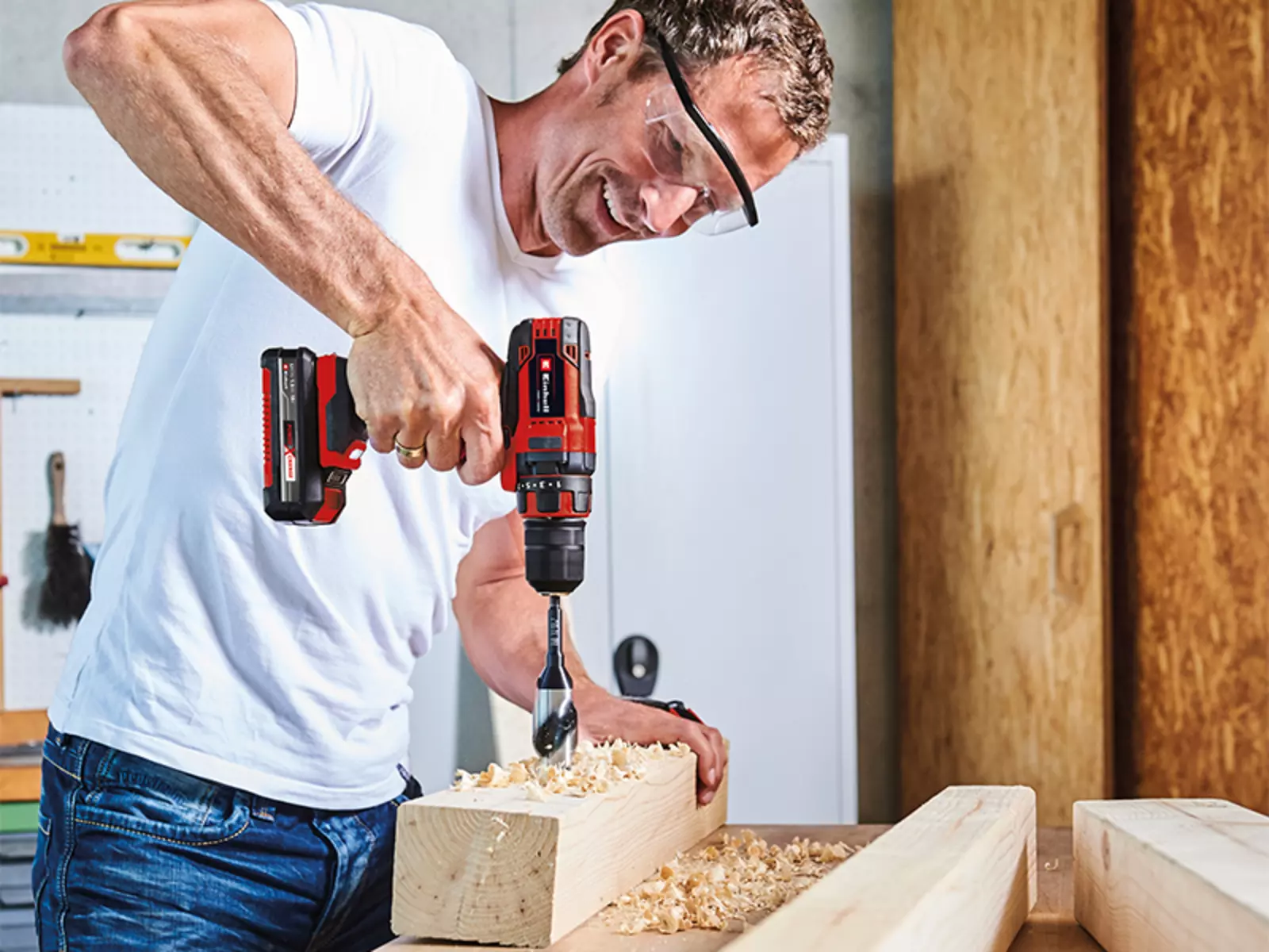 A man drills into a thick piece of wood on a workbench using the Einhell cordless drill driver TC-CD 18/35 Li, while wearing safety goggles.