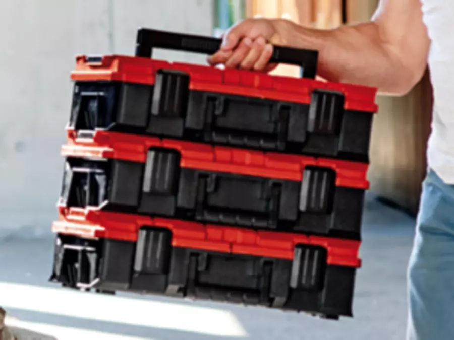 Man carrying three connected Einhell E-Case tool boxes with red lids using a foldable black handle.