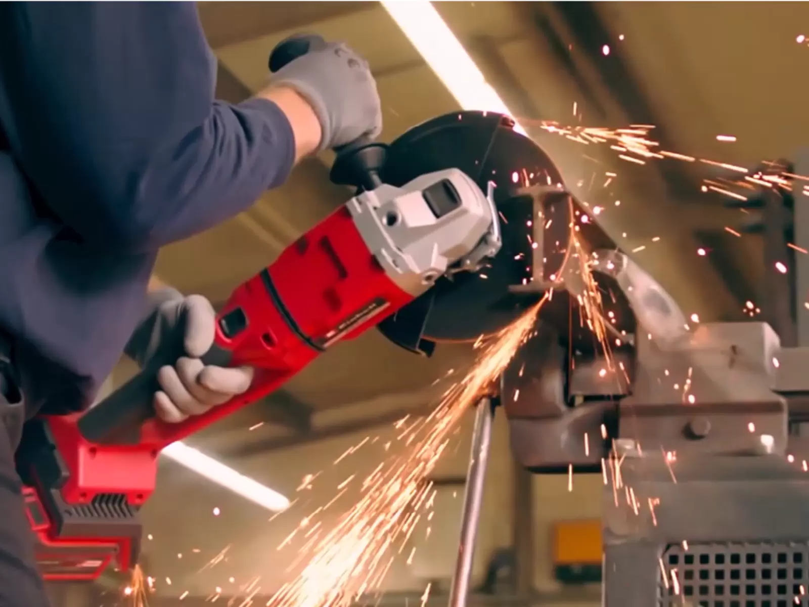 Seitenansicht eines Mannes, der mit beiden Händen einen großen, rot-schwarzen Akkuwinkelschleifer führt, um ein eingespanntes Metallteil zu trennen, während Funken in verschiedene Richtungen sprühen.
