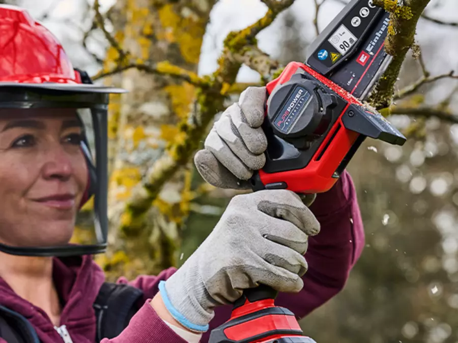 L’image montre une femme portant un casque et des gants qui coupe une branche à l’aide de l’élagueuse sans fil Einhell GE-PS 18/15 Li BL.