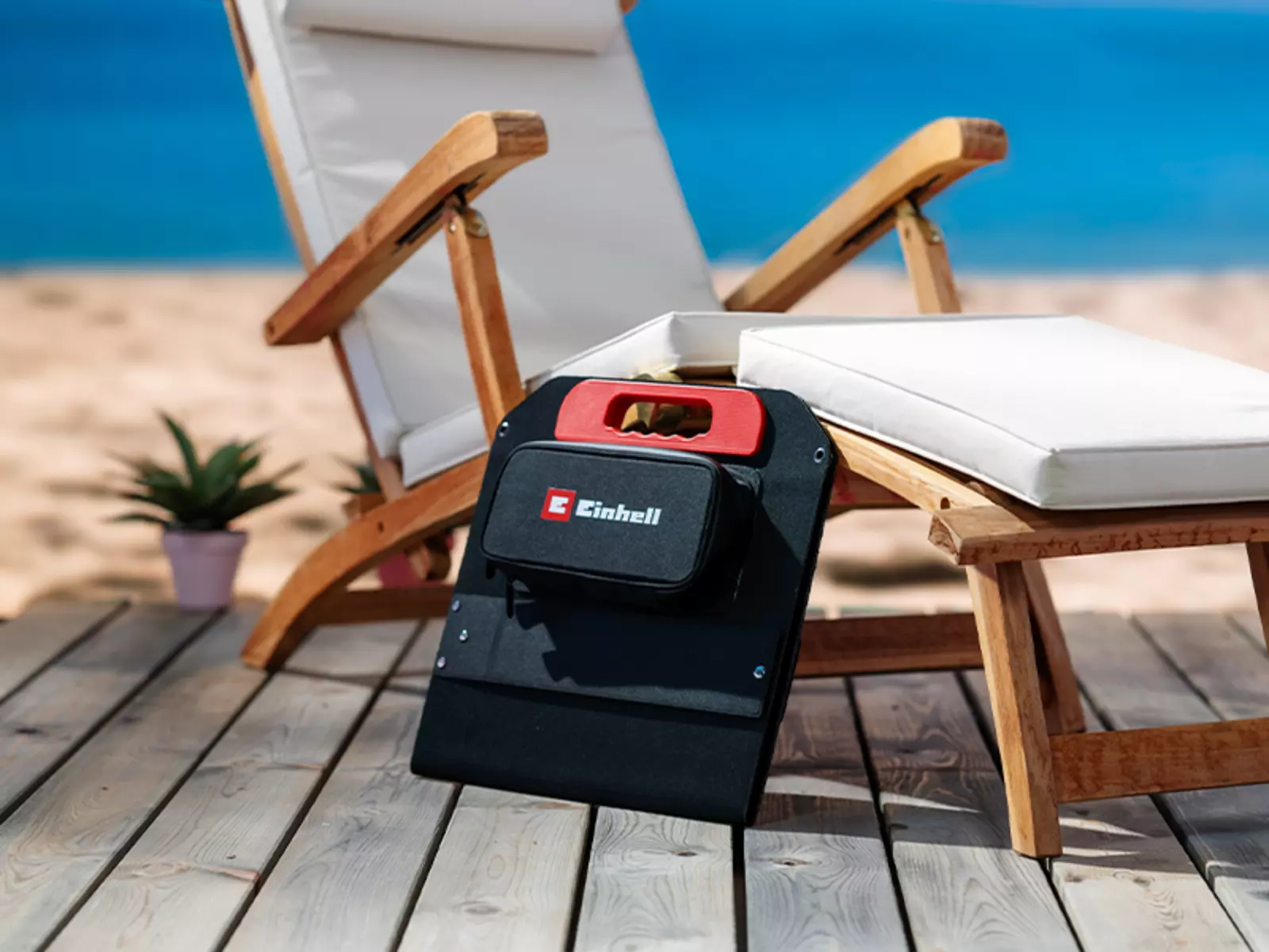 The folded Einhell solar panel with red carrying handle stands next to a sun lounger on a wooden deck at the beach.