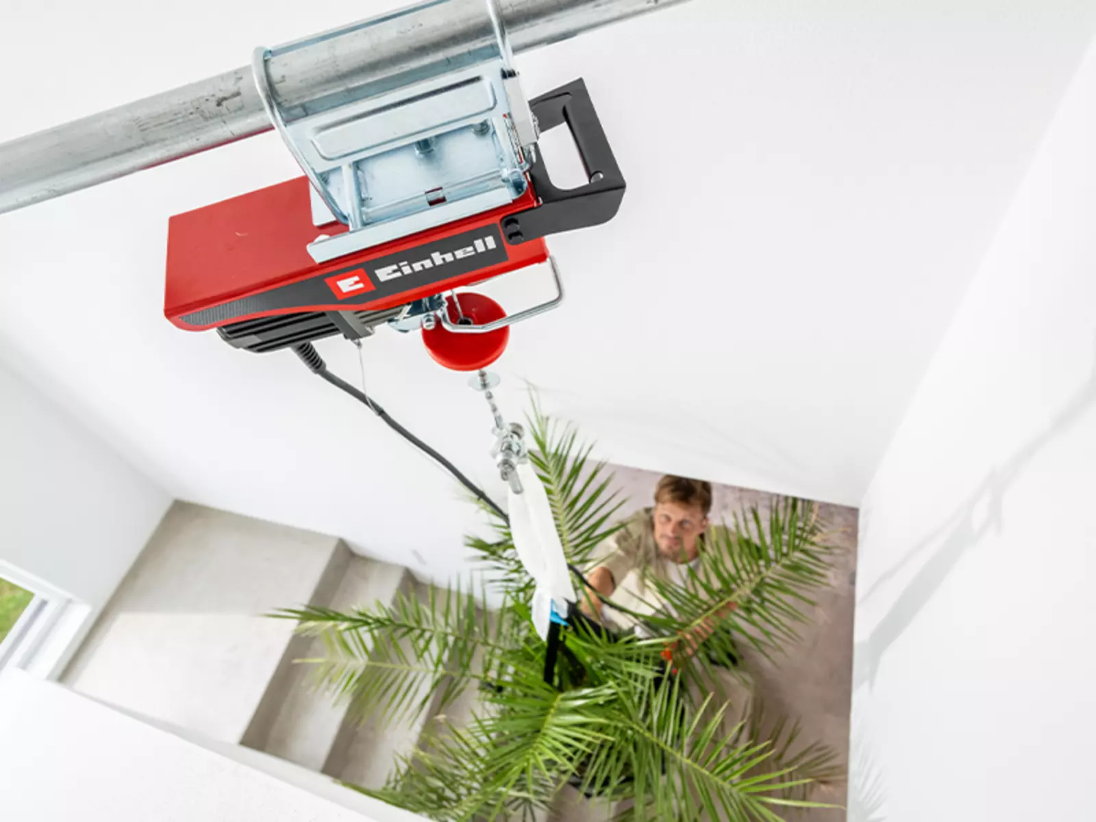 Einhell akülü halatlı vinç TE-EH 18/250 Li BL, bir merdiven boşluğunda bir bitkiyi kaldırırken bir adam aşağıdan yönlendirir.