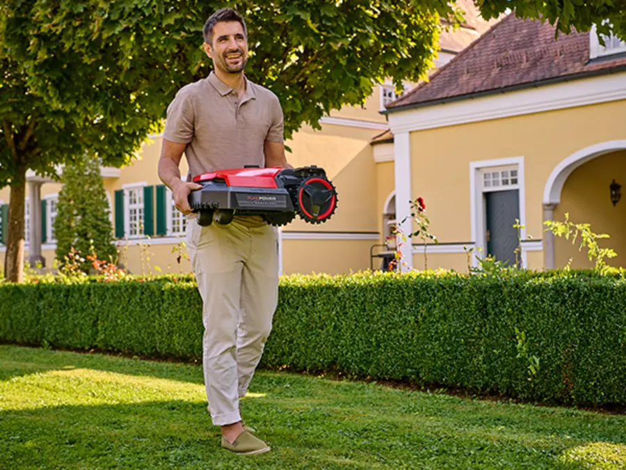 Un hombre lleva el Einhell robot cortacésped FREELEXO 1000 sobre un prado frente a un edificio de viviendas.