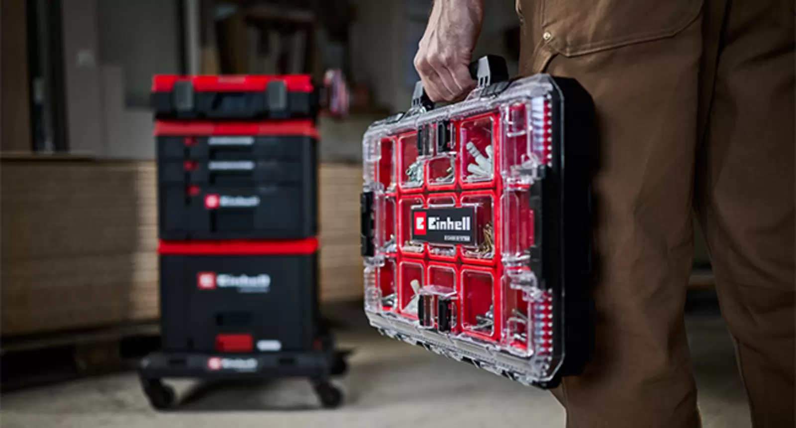 Una persona lleva un E-Case Einhell con tapa transparente; al fondo se ven E-Cases apilados sobre una base con ruedas.