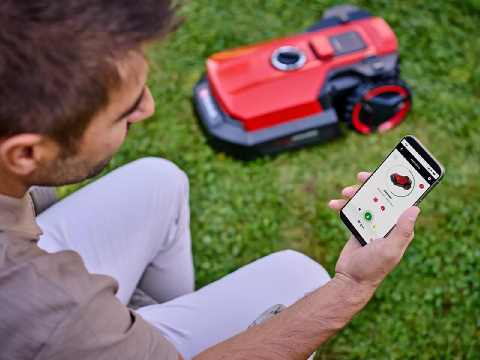 Un hombre se sienta en el césped y controla el Einhell robot cortacésped FREELEXO 1000 a través de una aplicación para teléfonos inteligentes; el robot rojo está en el fondo sobre la hierba.