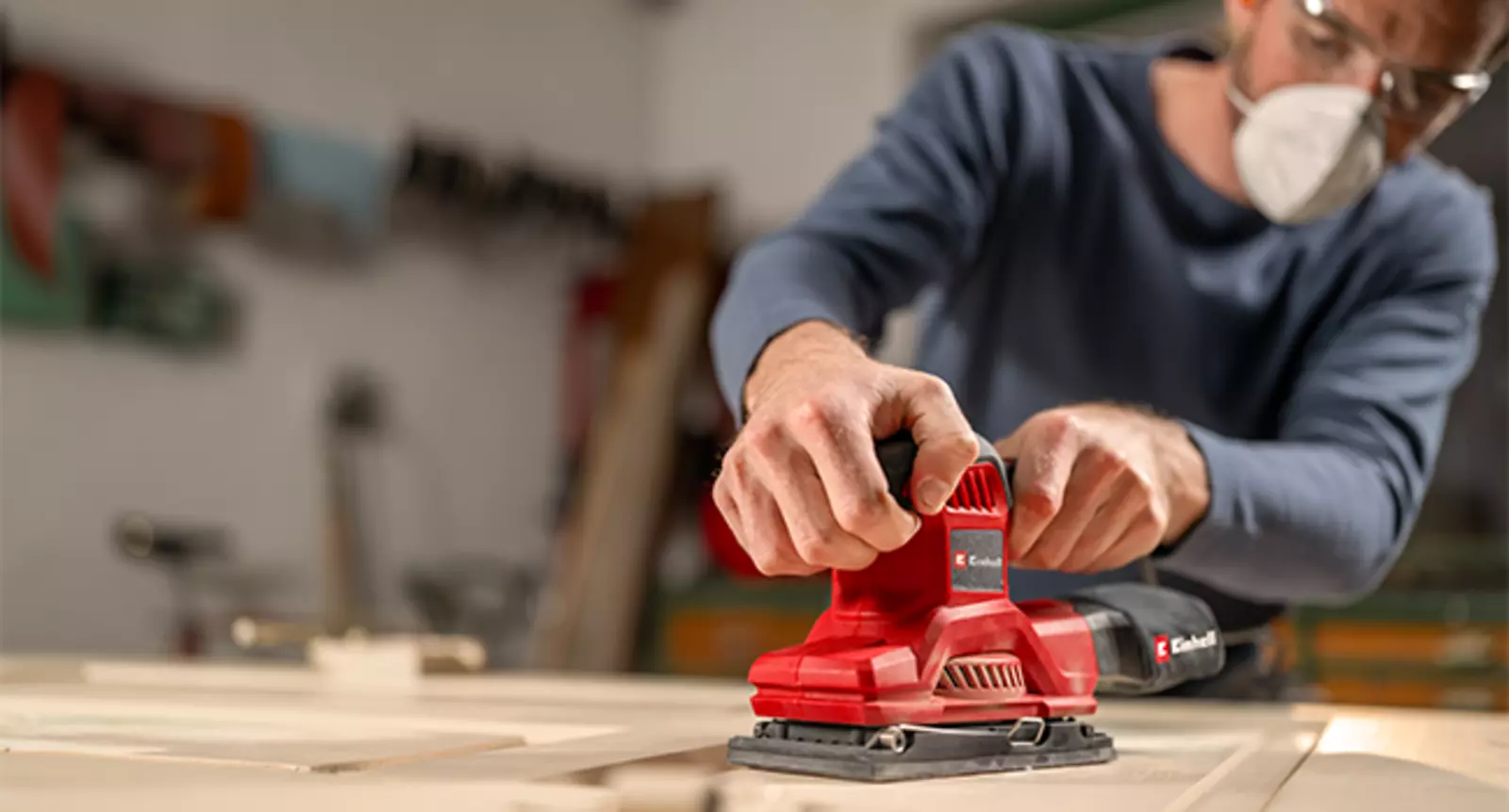 The Einhell orbital sander TC-OS 250 in red and black is used with a dust collection bag while sanding a wooden surface.
