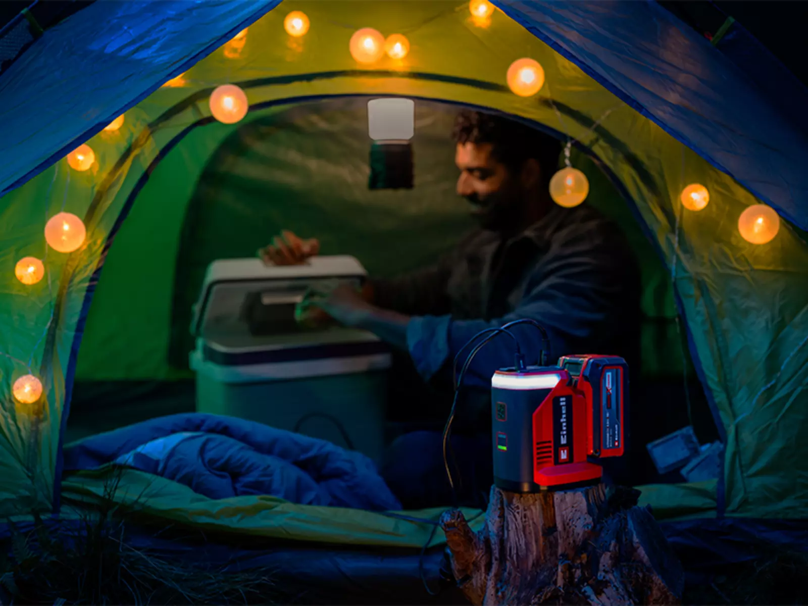 Einhell cordless energy station TE-ES 18/150/1 Li-C-Solo powering a string of lights inside a tent; in the background, a man sits in front of an open cooler.