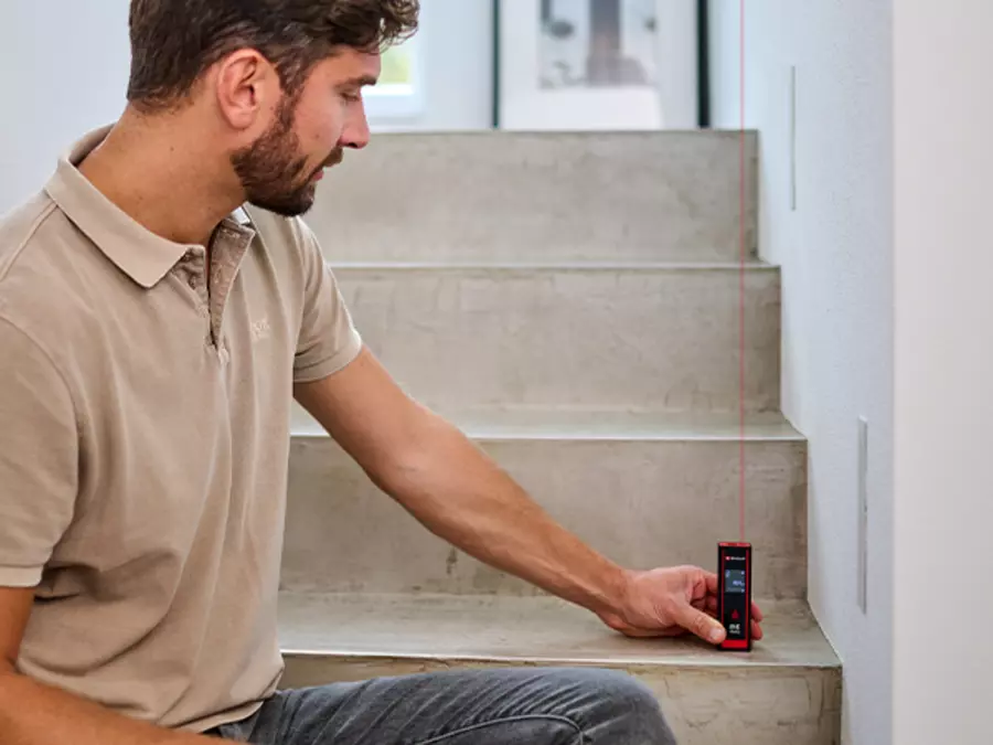 Um homem mede a área de uma escada de cimento com o Einhell medidor de distância a laser TC-LD 40 R; o feixe de laser vermelho é visível.