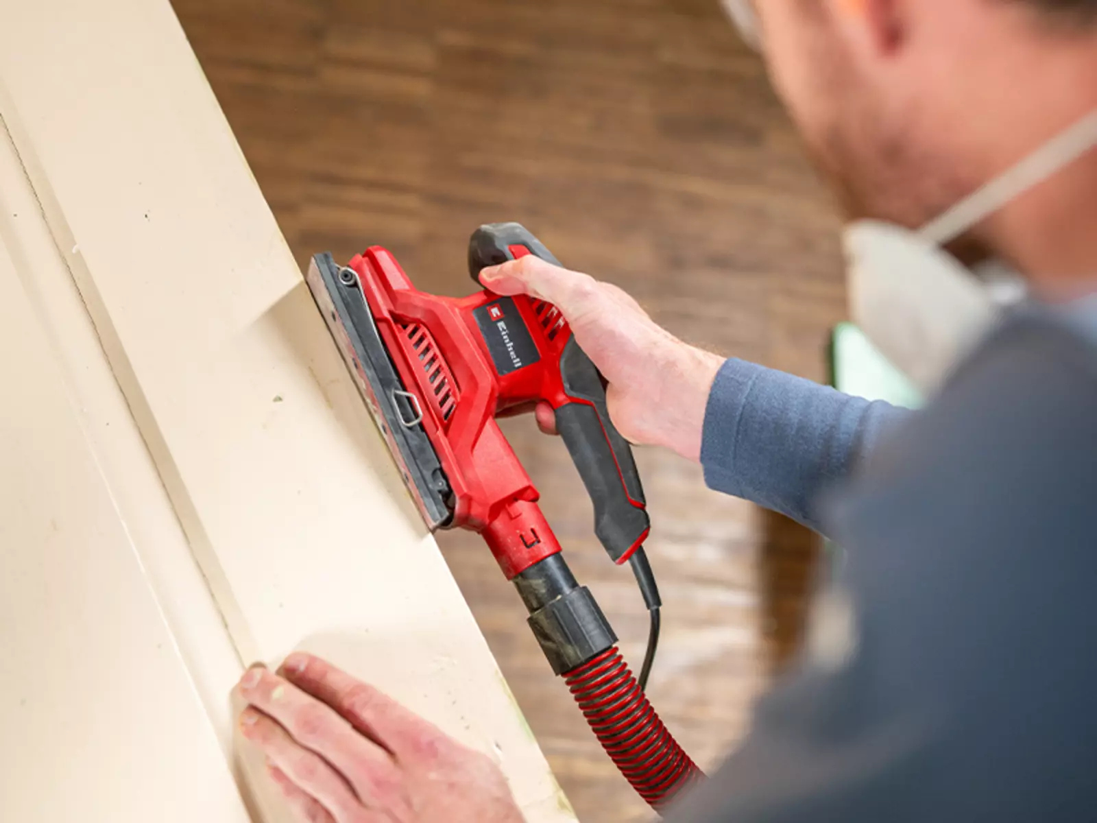 The Einhell orbital sander TC-OS 250 in red and black is held while sanding a wooden surface with a vacuum hose attached.