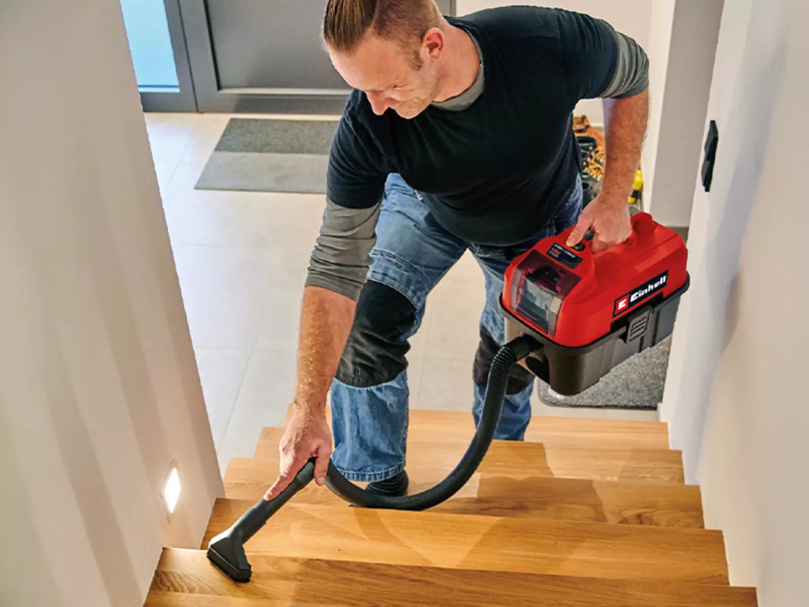 A man vacuums a staircase using the Einhell cordless wet and dry vacuum cleaner TE-VC 18/10 Li while carrying it by hand.