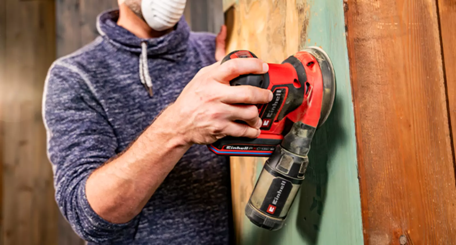 A user wearing a respiratory mask uses the Einhell cordless orbital sander TE-RS 18/28 Li - Solo with a dust collection box on a vertical wooden surface.