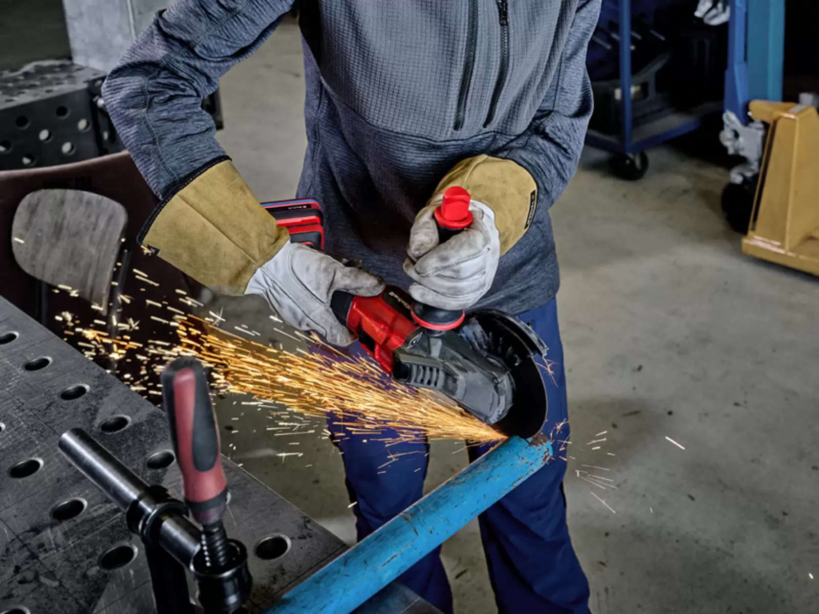 Close-up of a person’s hands wearing protective gloves cutting a clamped, blue-painted metal pipe with an angle grinder, sparks fly from the workpiece.