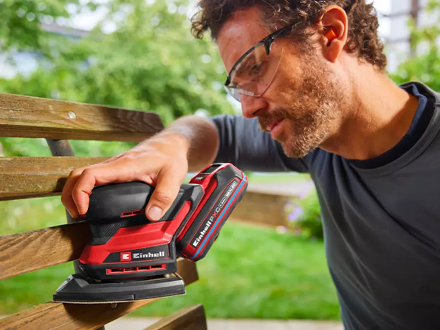 The Einhell cordless multi sander TE-OS 18/150-2 Li is used by a person outdoors to sand wood.