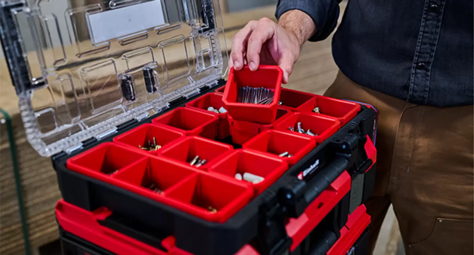 Una mano levanta un compartimento rojo con piezas pequeñas de un E-Case Einhell abierto; los demás compartimentos contienen accesorios.