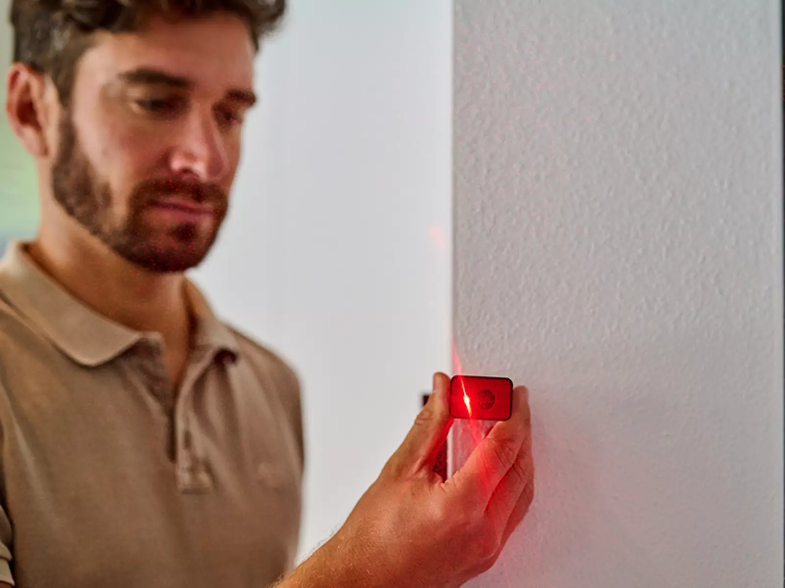 Um homem segura o Einhell medidor de distância a laser TC-LD 40 R contra uma parede branca; o feixe de laser vermelho é ativado.