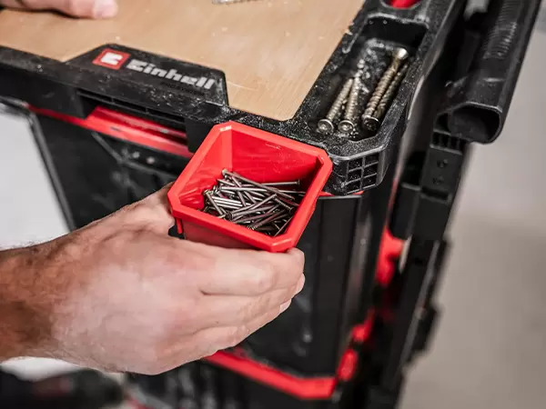 Red Modular Box filled with nails being placed into the integrated storage compartment of the Einhell Pro System Work Table.
