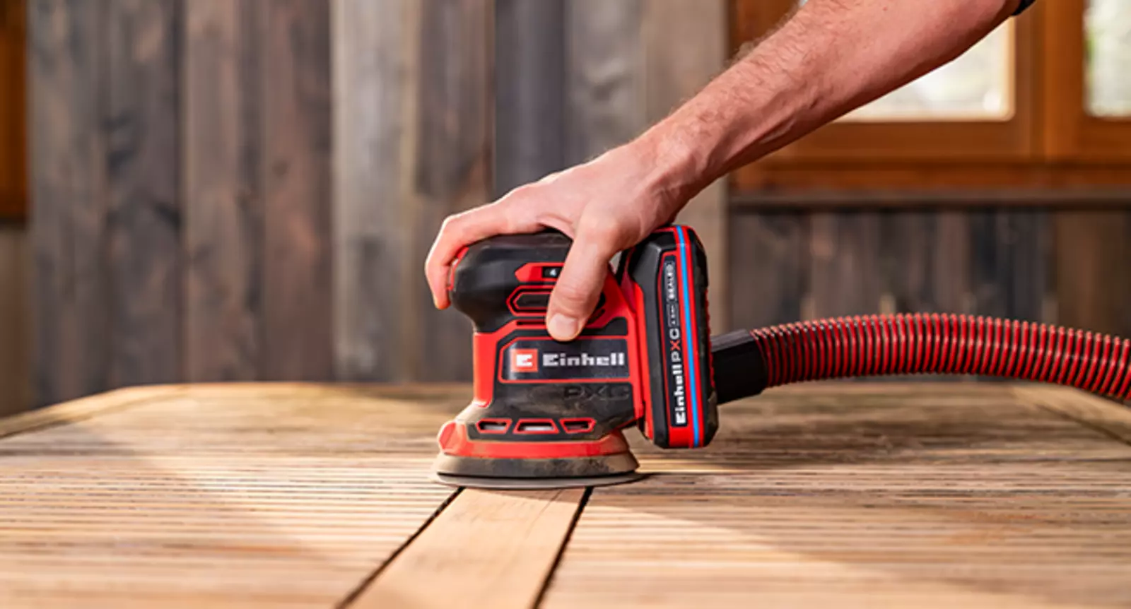 The Einhell cordless orbital sander TE-RS 18/28 Li - Solo is used with a vacuum hose attachment to sand a wooden table.