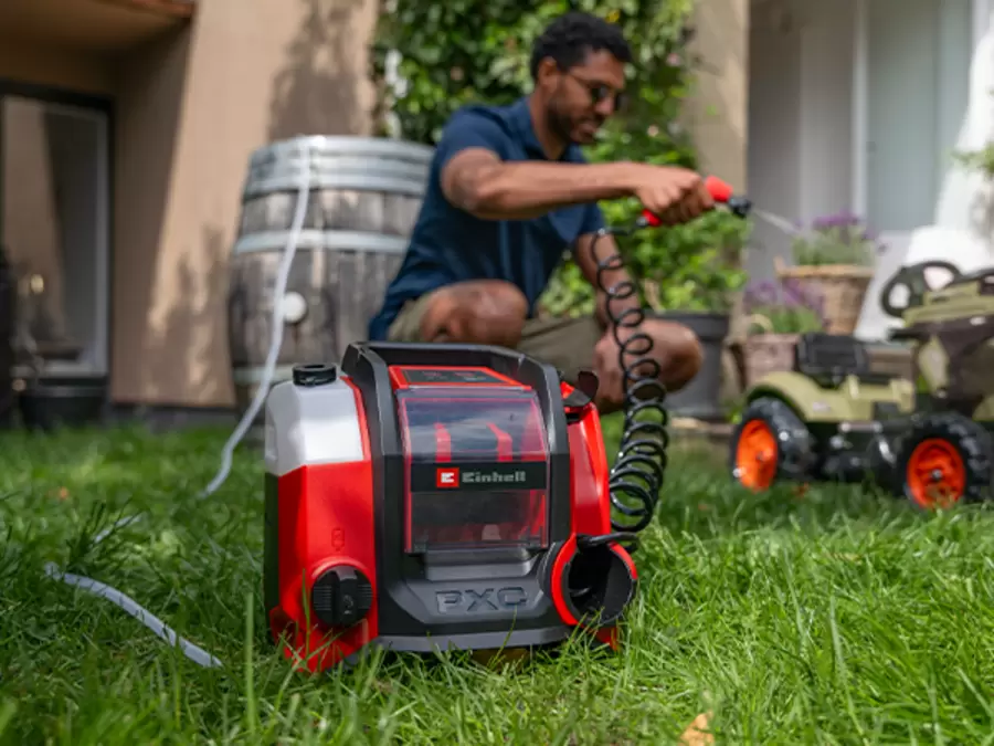 The Einhell cordless pressure cleaner TE-HP 18/10 Li D-Solo stands in a garden and draws water from a water butt through a suction hose.