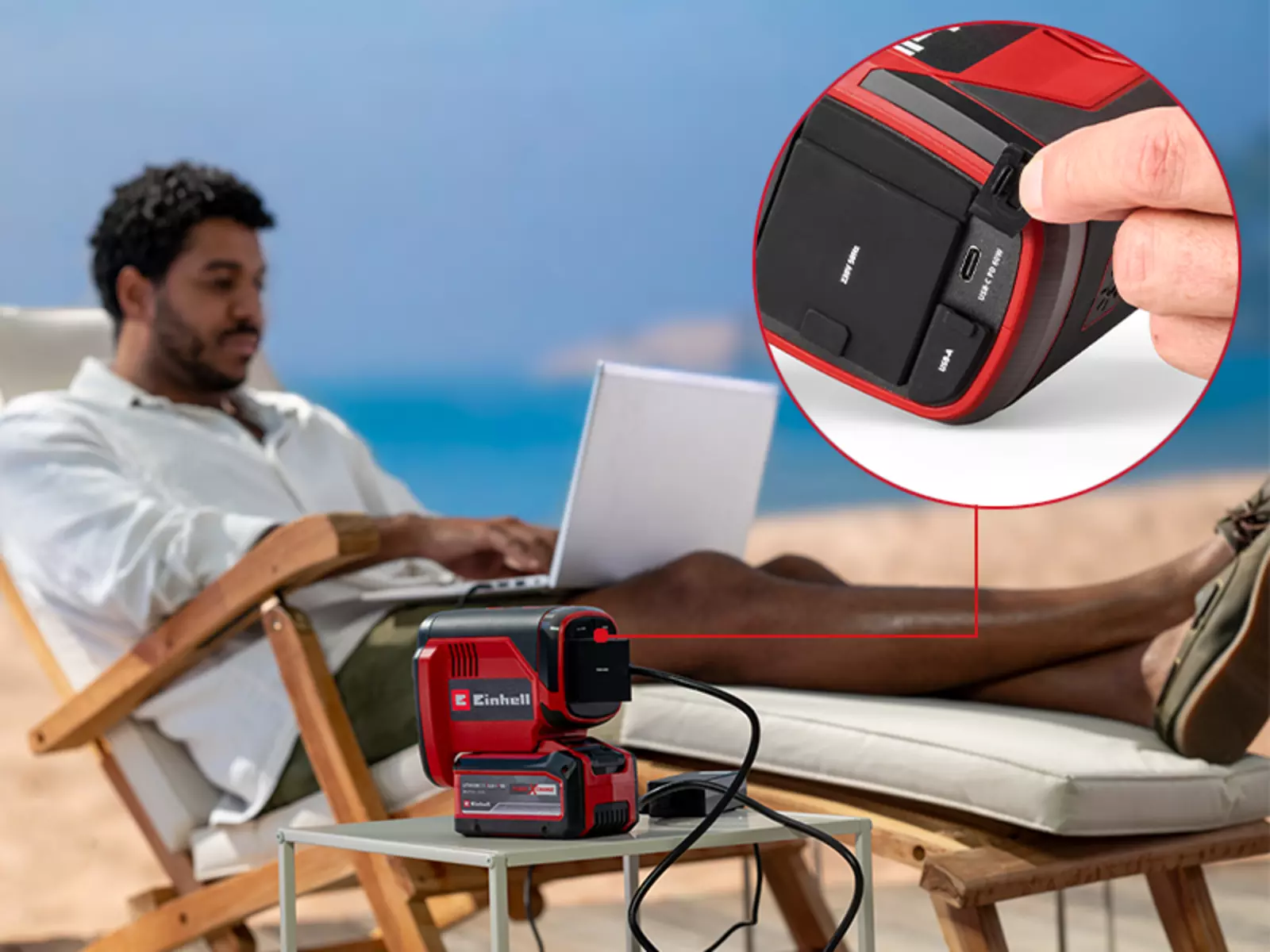Ein Mann sitzt am Strand mit einem Laptop, der über die Einhell Akku-Energiestation TC-ES 18/150/1 Li-Solo mit Strom versorgt wird; im Detailausschnitt ist ein Finger zu sehen, der auf die USB-C-Power-Delivery-Buchse der Energiestation zeigt.