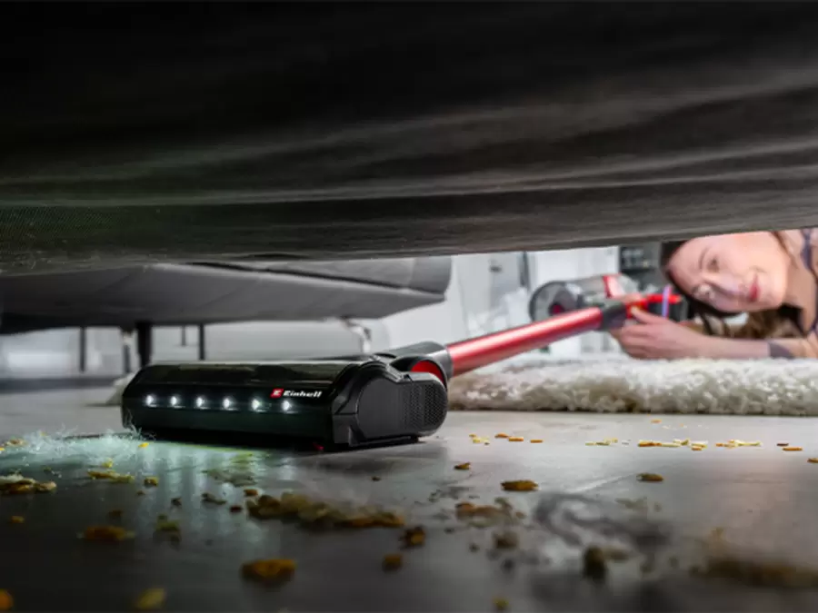 A person uses the Einhell cordless stick vacuum cleaner TE-SV 18/270/1 Li BL-Solo to clean the floor under a low piece of furniture.